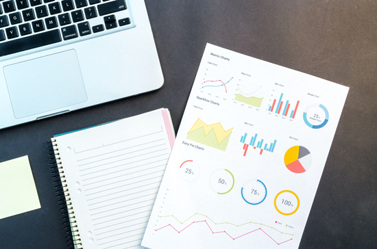 Laptop on a desk with some printed sheets containing graphs and charts.