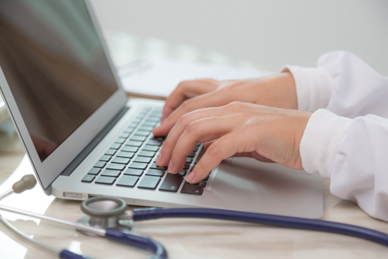 A close up of a man's hands using a laptop keyboard with a stethoscope next to it.