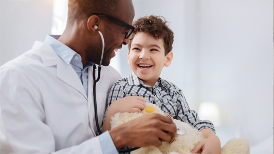 Doctor holding a stethoscope against a soft toy bear whilst a child looks on and laughs.