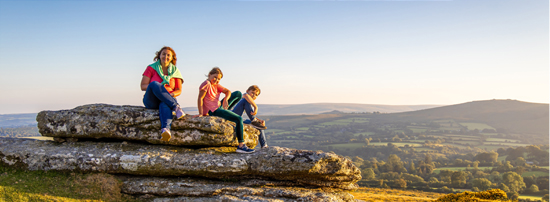 A woman sat on a large rock in the countryside with her two children.