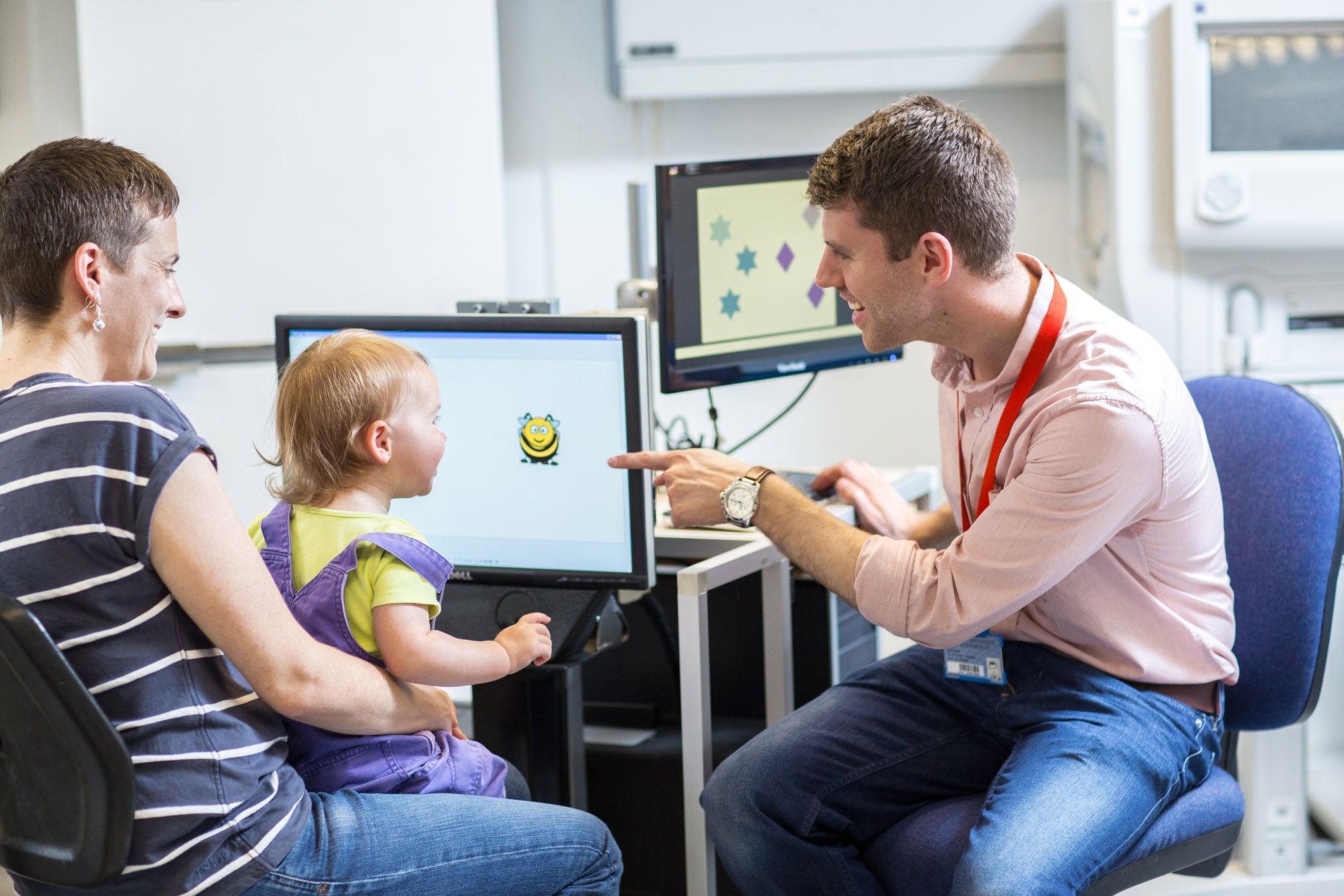 A happy mother, child and doctor sat at a computer. The doctor is pointing at an drawing of a bumble bee on the screen.