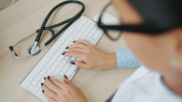 Clinician in glasses typing - image focus on keyboard with stethoscope beside it